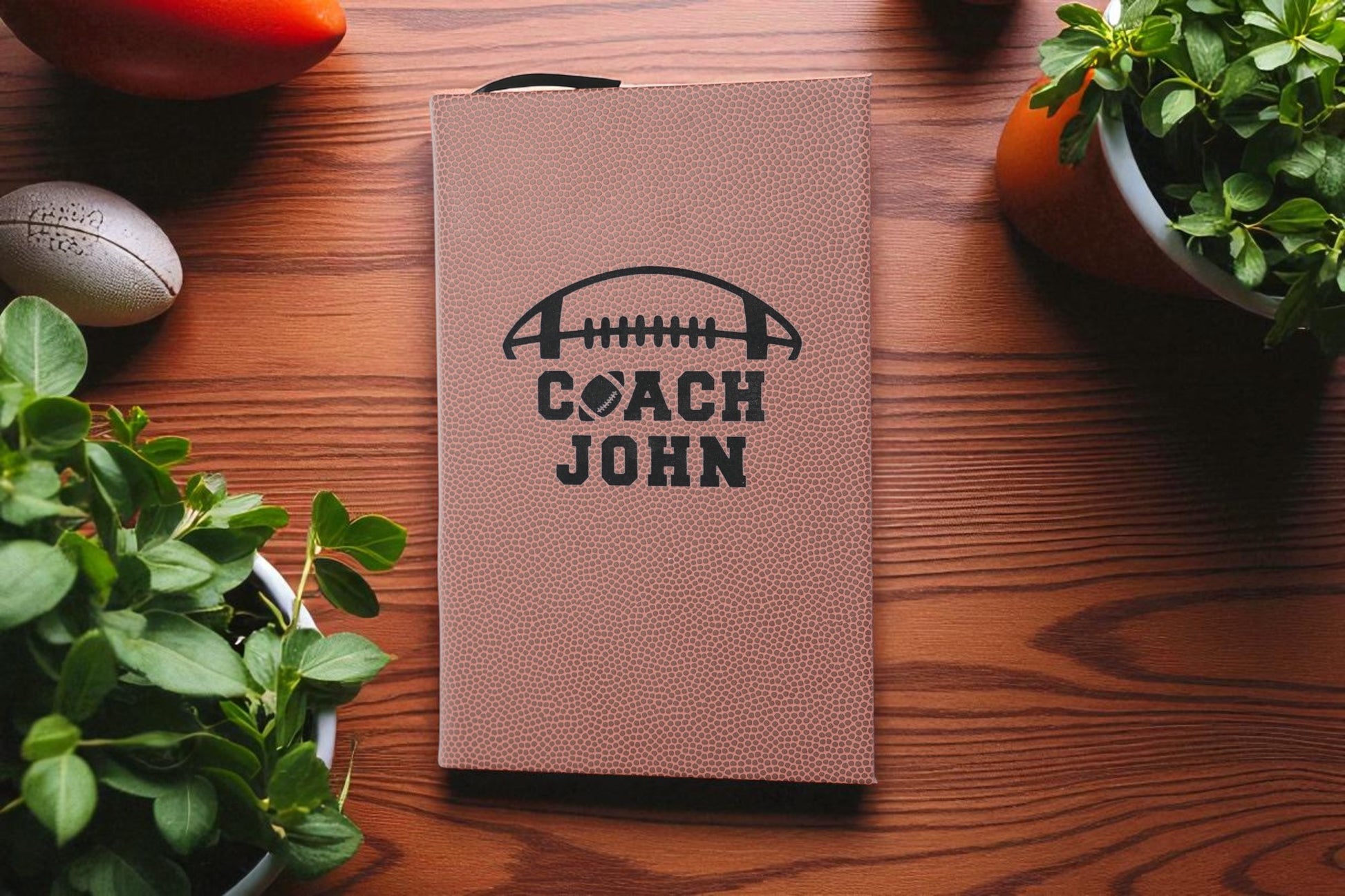 Brown leather notebook with 'Coach John' engraving on a wooden surface with plants.