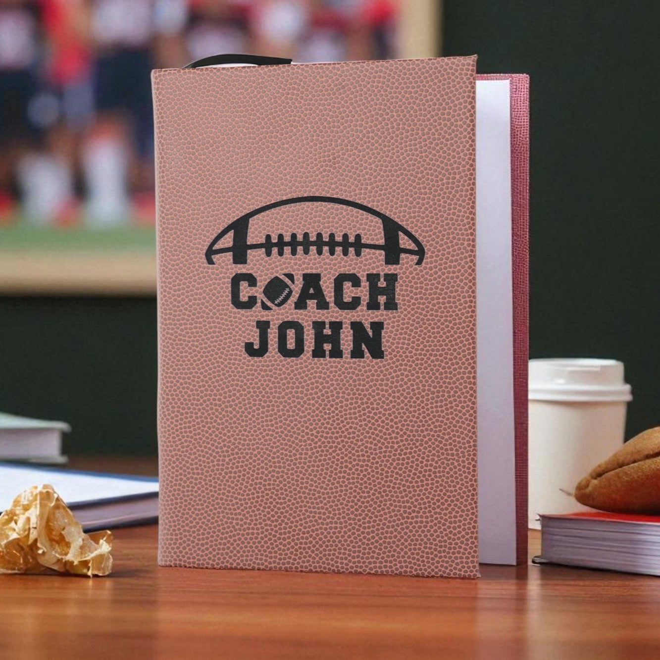 Notebook with 'Coach John' on a desk with a blurred sports event background