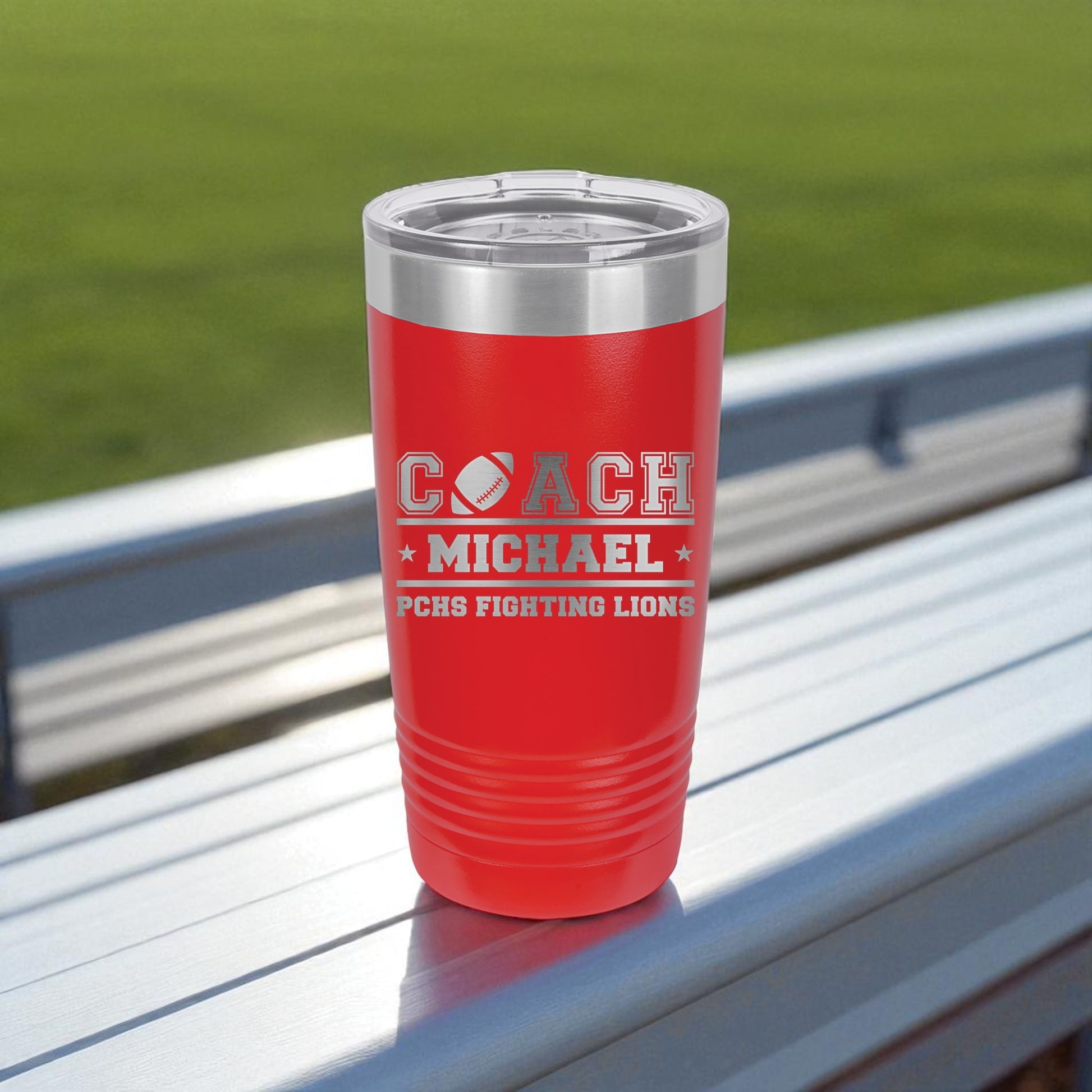 Red insulated tumbler with 'COACH MICHAEL' text on a metal surface with grass background