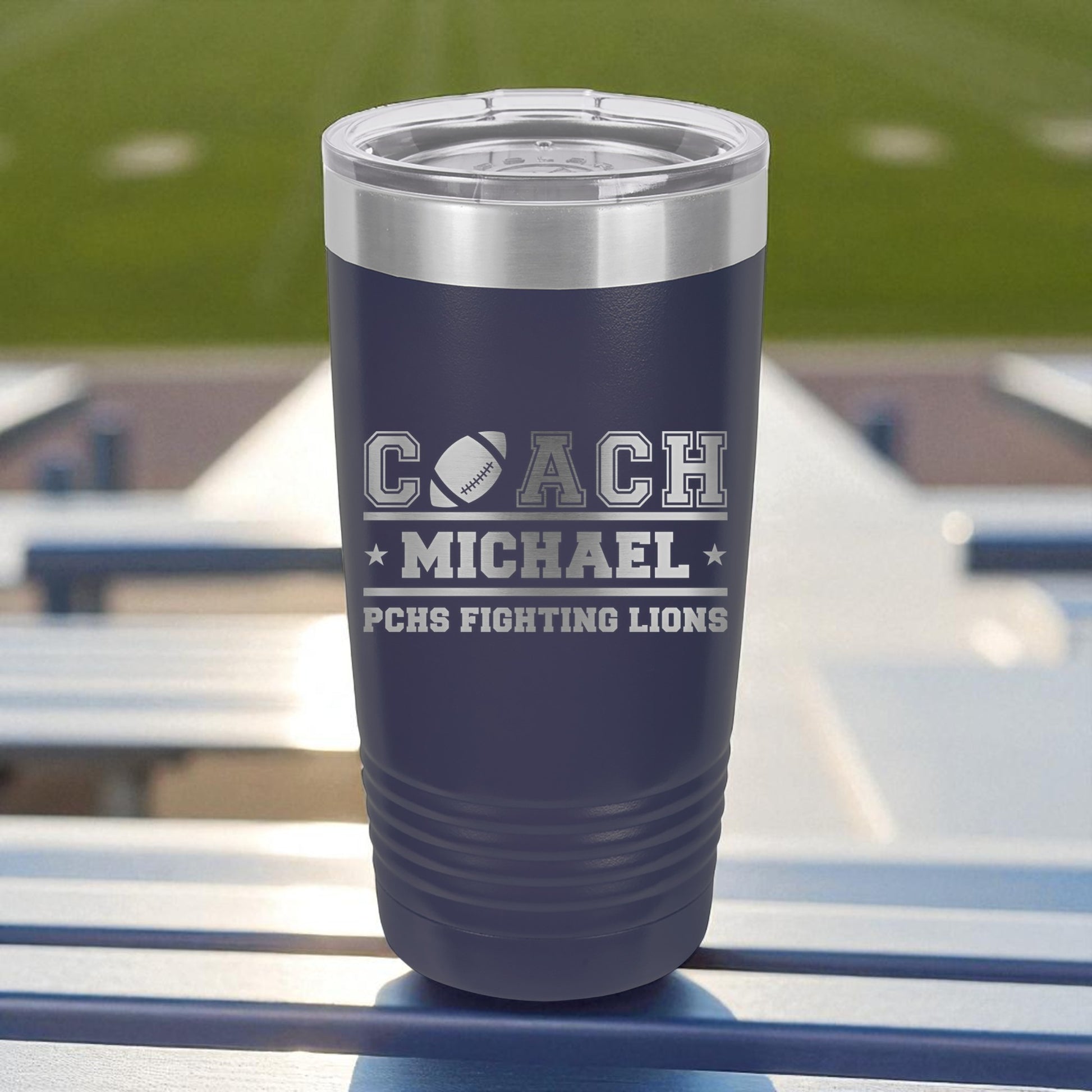 Navy blue tumbler with engraved text on a metal bleacher at a sports field.