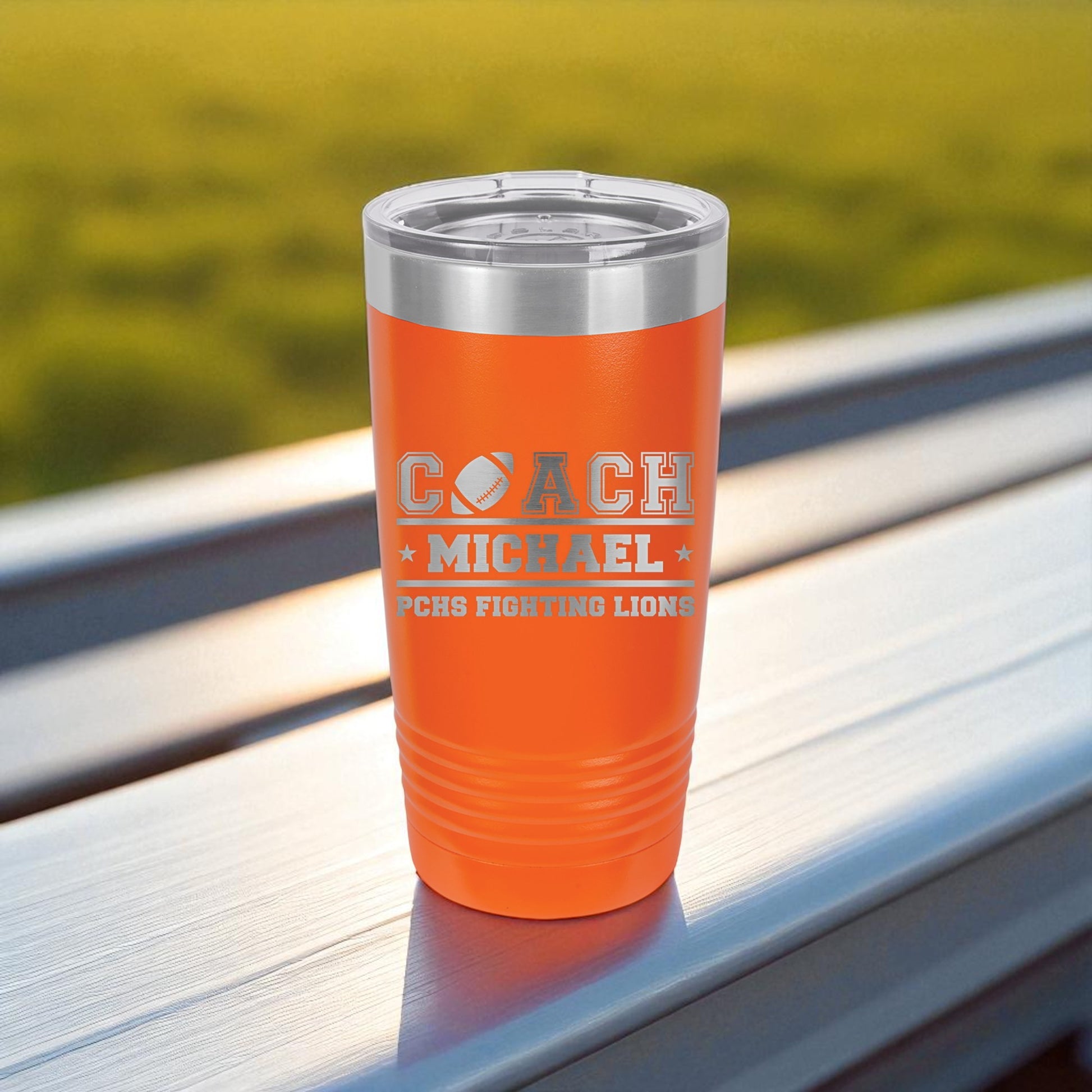Orange tumbler with 'COACH MICHAEL' engraving on a wooden surface with a blurred green background