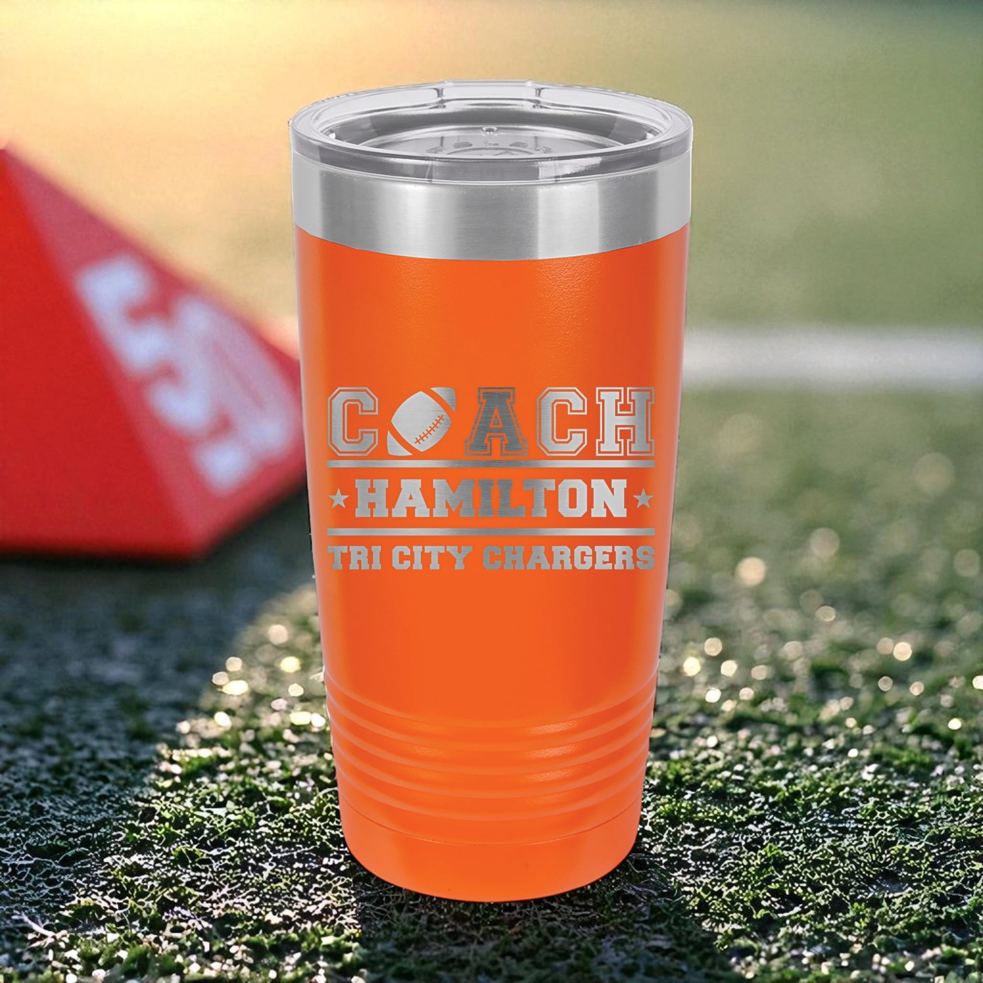 Orange tumbler with 'Coach Hamilton Tri City Chargers' text on grass