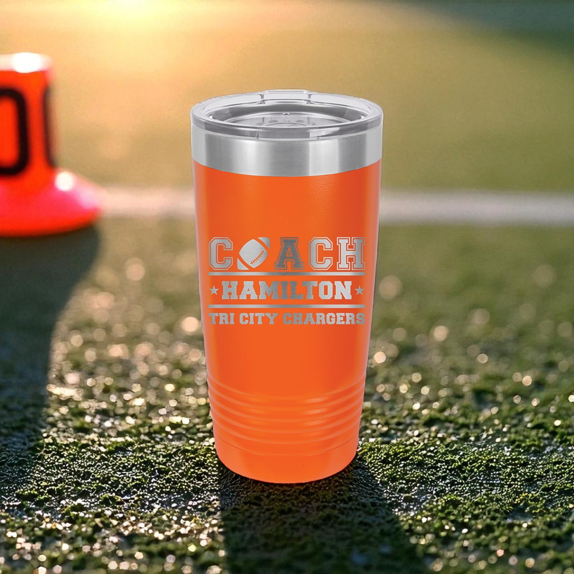 Orange tumbler with 'COACH HAMILTON TRI CITY CHARGERS' text on a grassy field background