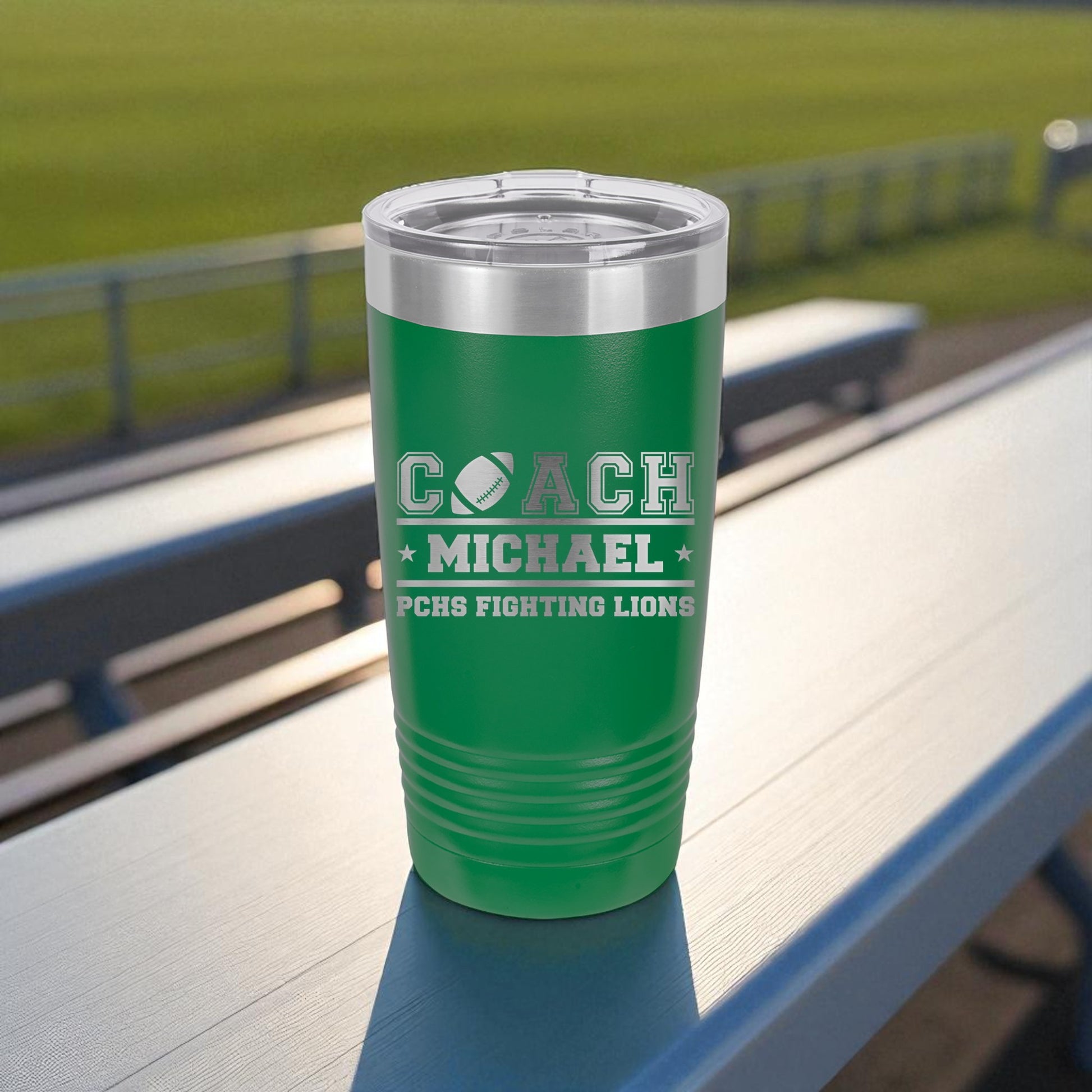 Green tumbler with 'COACH MICHAEL' and 'PCHS FIGHTING LIONS' text on a table outdoors.
