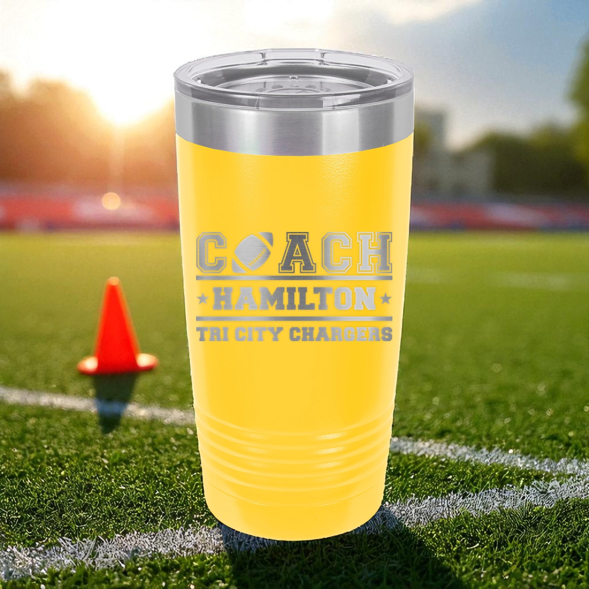 Yellow tumbler with 'COACH HAMILTON THE CITY CHARGERS' text on a sports field with a cone.