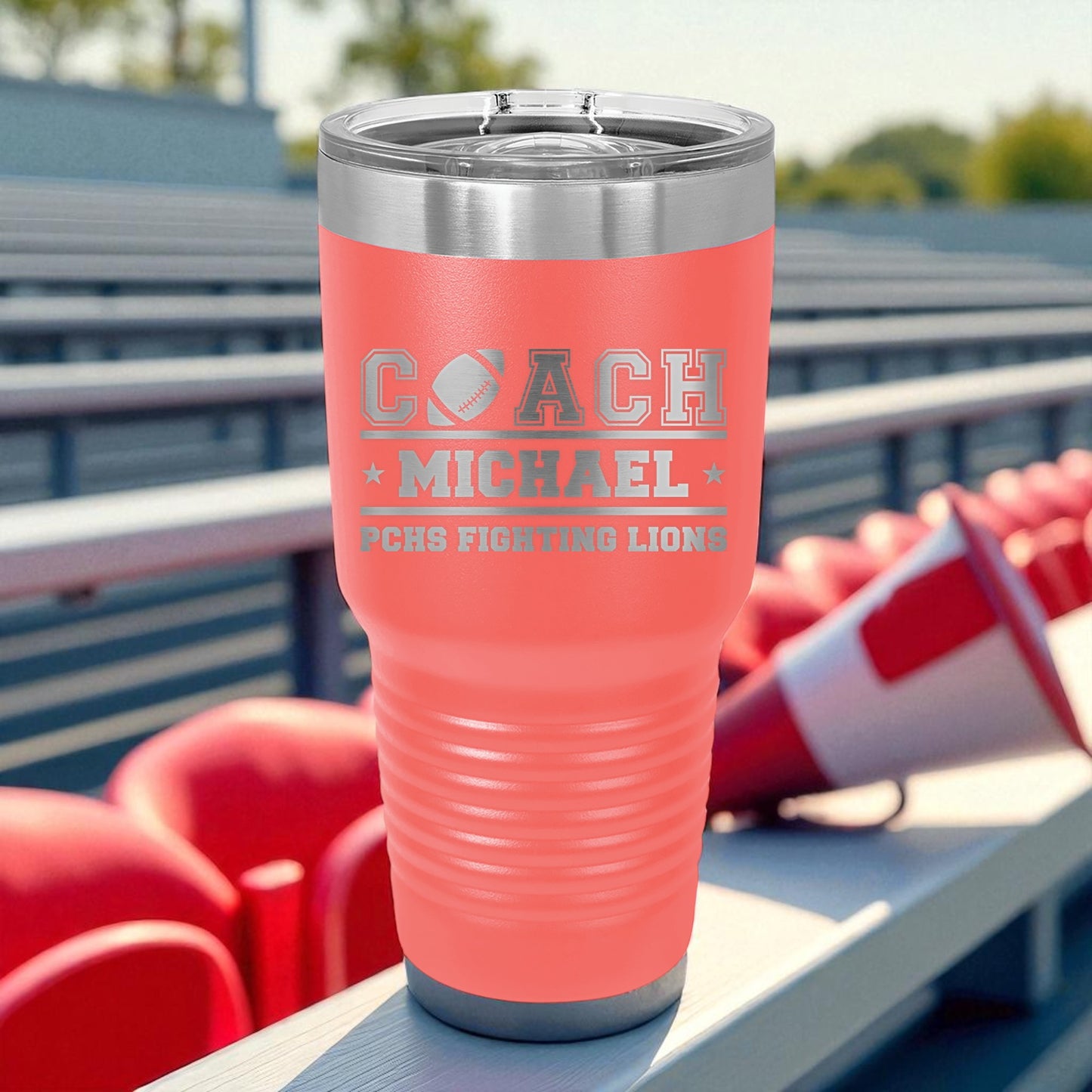 Pink tumbler with 'COACH MICHAEL' and 'PCNS FIGHTING LIONS' text, placed on bleachers.
