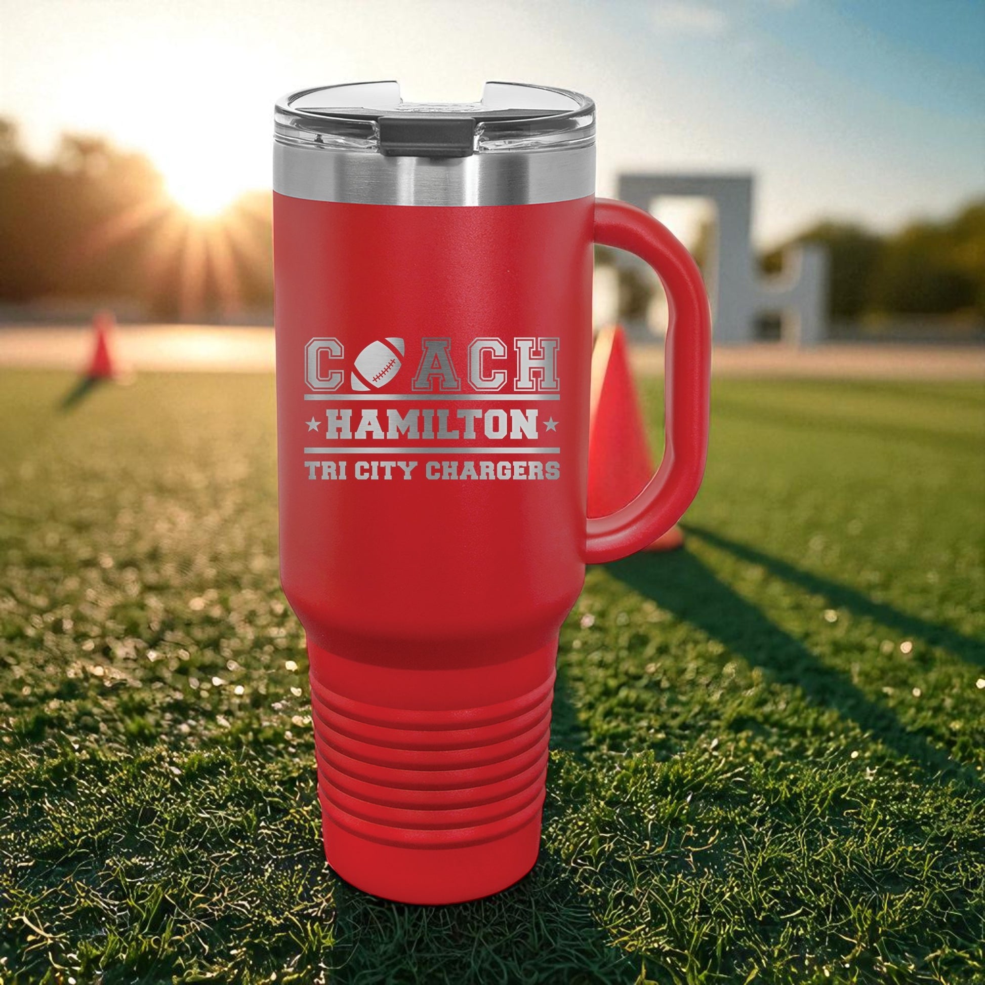 Red travel mug with 'Coach Hamilton' text on grass field