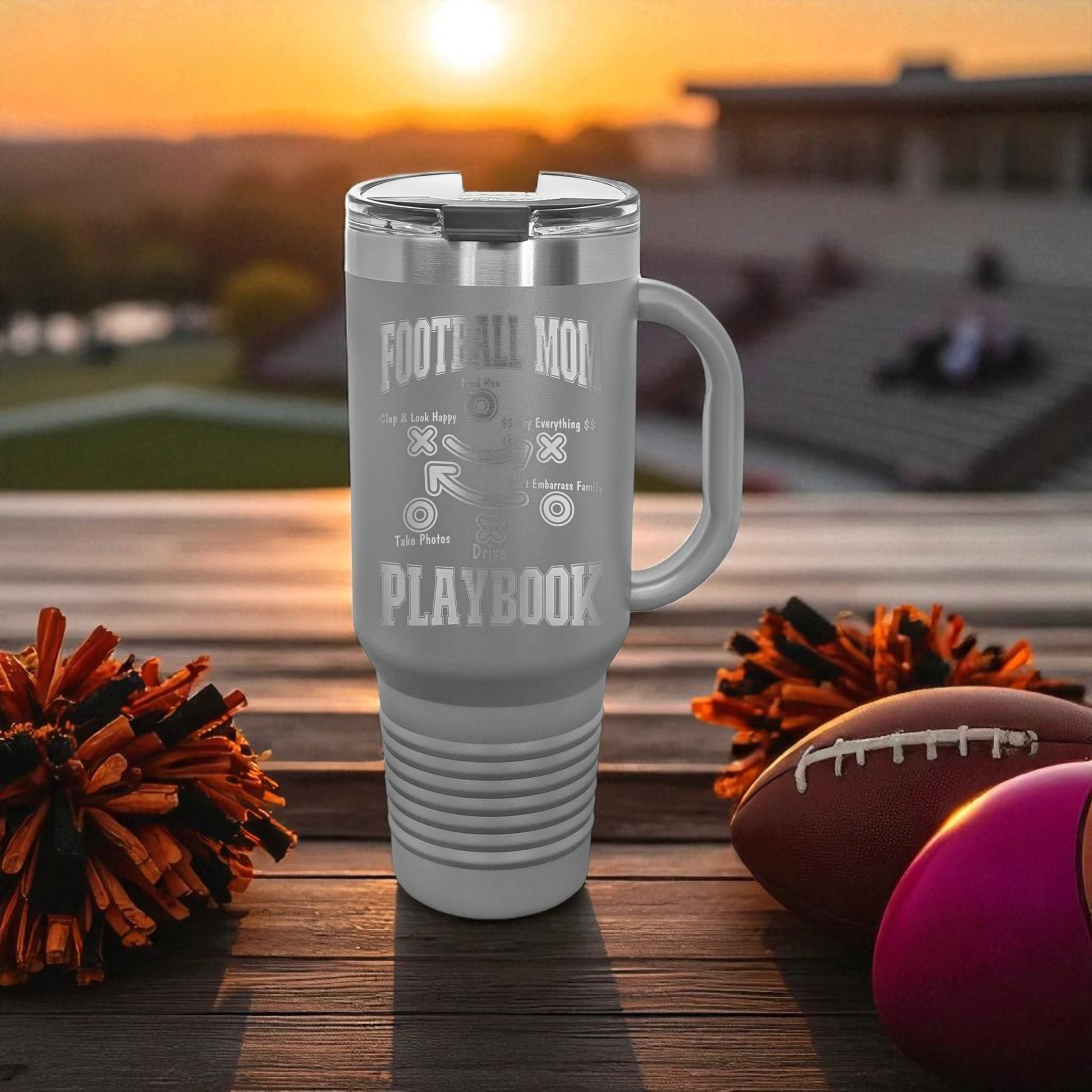 Gray travel mug with 'FOOTBALL PLAYBOOK' text on a wooden surface with sports-themed items and sunset background.