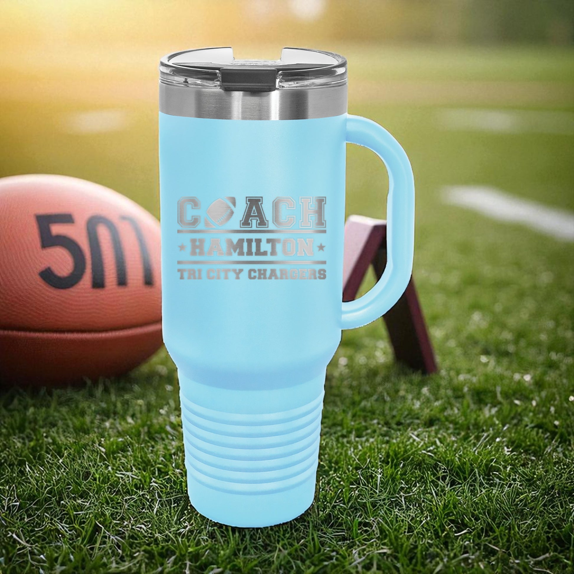 Light blue travel mug with engraved text on a grassy field with a football and goal post in the background.