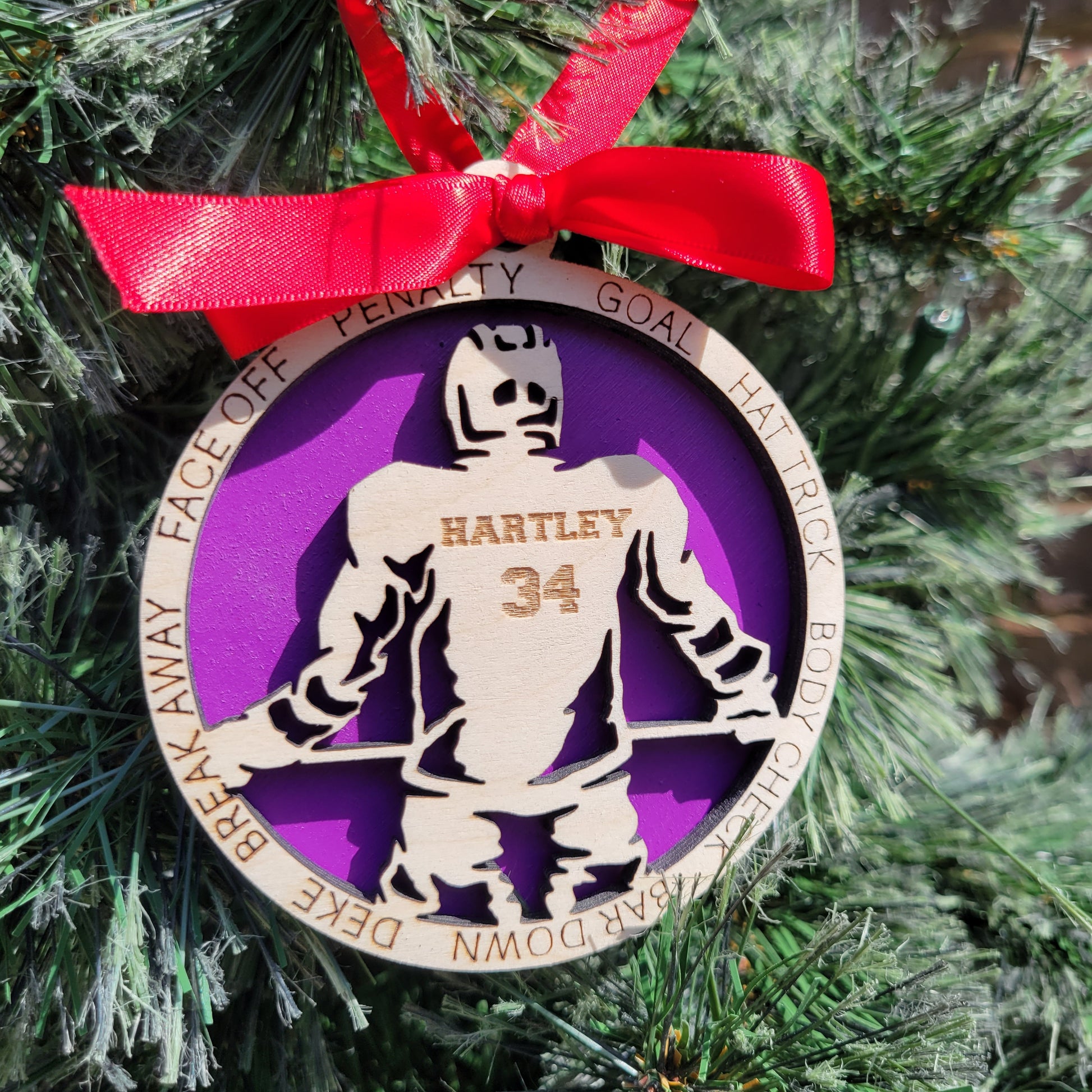 Decorative ornament with a Decorative Christmas ornament with a hockey player, personalized hockey player ornament, custom wooden hockey player ornament, hockey player ornament, christmas gift for hockey player on a purple background, attached to a Christmas tree with a red ribbon.