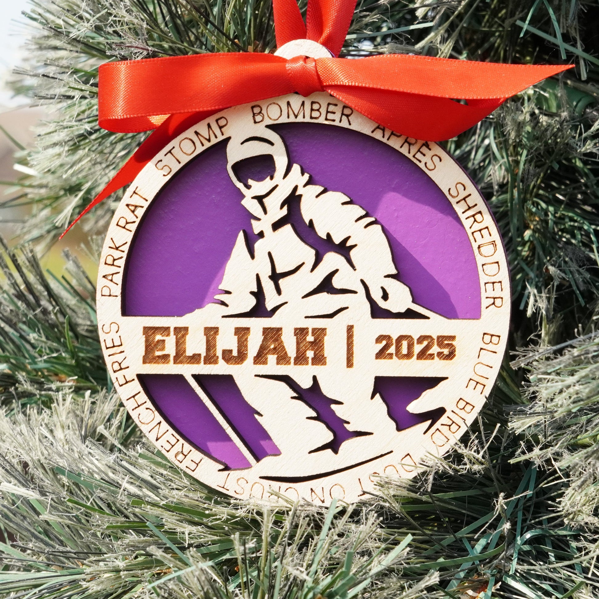 Decorative ornament with a purple and gold design, featuring the name 'Elijah' and the year '2025', hanging on a Christmas tree. skier ornament, skiing ornament