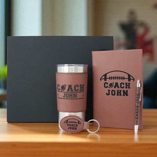 Coach John branded tumbler, notebook, and keychain on a wooden surface with a blurred background.