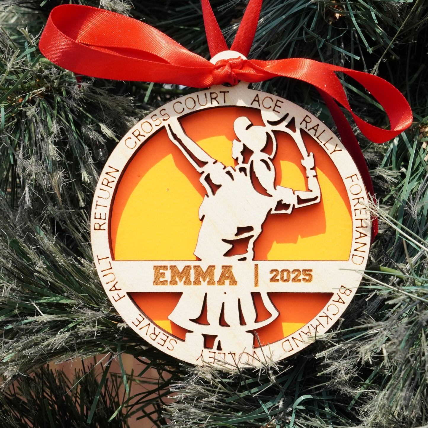 Decorative ornament with a tennis player design and 'EMMA 2023' text, tied with a red ribbon on a Christmas tree.